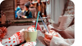 Hands hold hot cocoa in front of TV showing a holiday movie.
